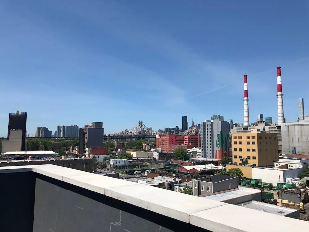 A view over rooftops and buildings toward the Queensboro Bridge and two red-and-white smokestacks, taken from a raised terrace with a concrete ledge in the foreground.