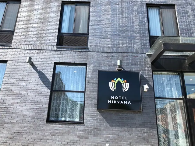 Exterior of a grey brick building with a black sign reading Hotel Nirvana, windows with sheer curtains, and a glass canopy over the entrance.