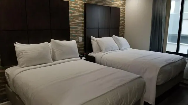 Two queen beds with white bedding and dark padded headboards sit in a room with patterned wallpaper, beside a window looking onto a neighbouring building.