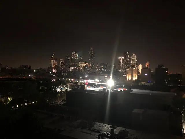 Night view from an elevated position over rooftops and a parking lot toward a skyline of lit high-rise towers.