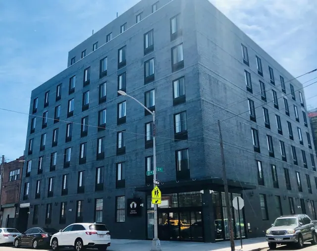 Exterior of a multi-storey dark brick hotel building on a street corner, with parked cars and a pedestrian crossing sign in front.