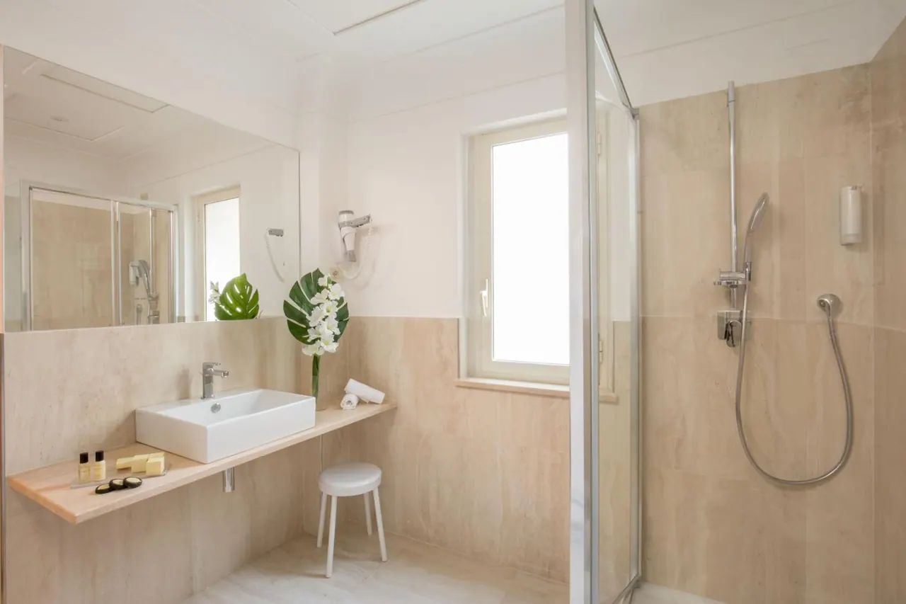 A bathroom with a single rectangular basin on a wooden shelf, a mirror, a window, a white stool and a walk-in shower with a glass door.