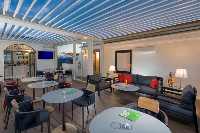 A covered terrace with a blue slatted pergola roof, grey tables and navy chairs, and a lounge sofa set with red cushions.