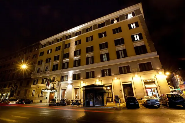 Night exterior of a yellow multi-storey hotel building on a Rome street, with parked cars and a kiosk in front.