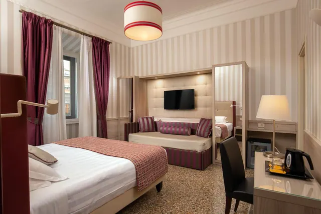 A hotel bedroom with a double bed, a sofa bed against a striped wall, a wall-mounted television, a desk with a kettle, and a small fridge under the desk.