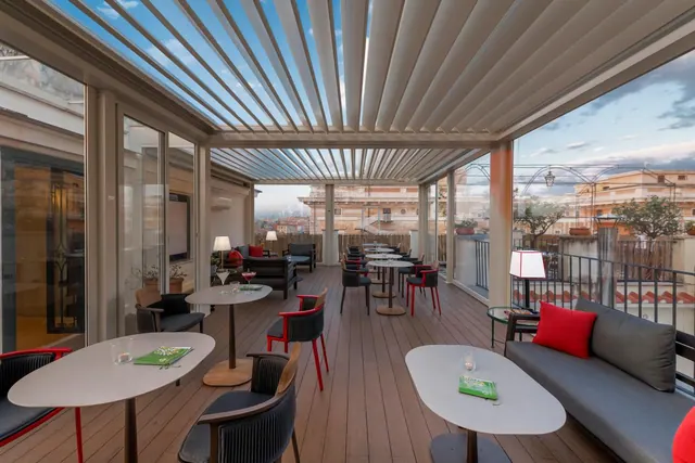 A covered rooftop terrace with tables, chairs and a grey sofa, with glass walls looking out over neighbouring rooftops.