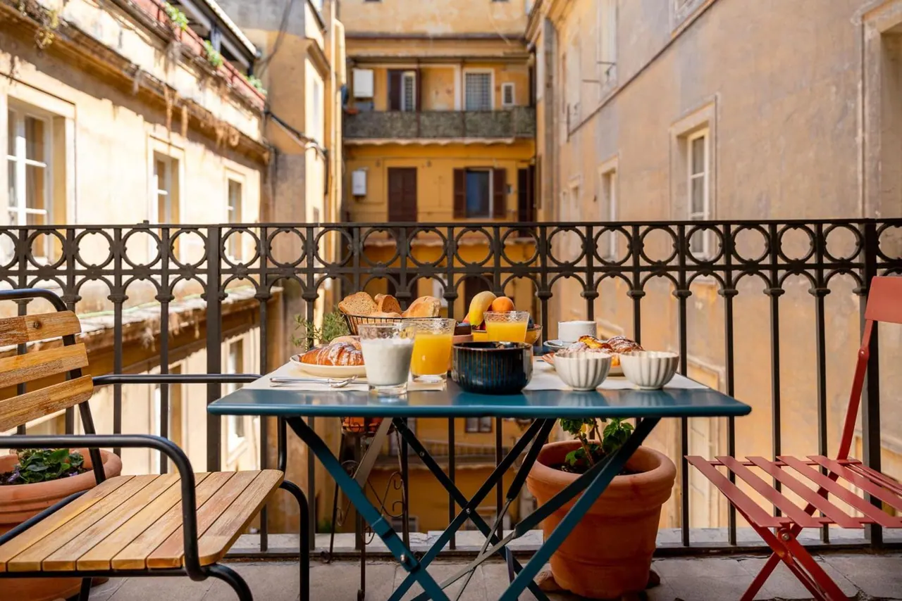 A breakfast table with bread, juice, fruit and bowls sits on a balcony overlooking a courtyard of old ochre buildings.