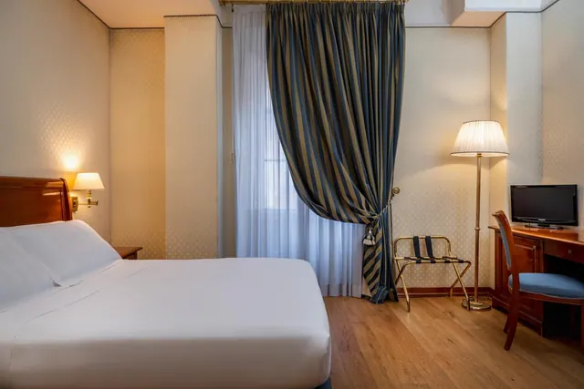 A bedroom with a double bed with white linen, a striped curtain by the window, a wooden desk with a chair, and a flat-screen television.