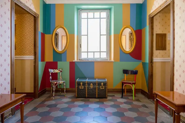 A symmetrical room with painted multicolour geometric stripes on the walls, two oval gold mirrors, a barred window, a patterned leather trunk and two chairs on a hexagonal tiled floor.