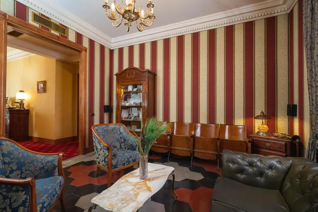 A lounge with red and cream striped wallpaper, a chandelier, blue upholstered armchairs, a leather sofa, a glass-fronted cabinet and a marble coffee table.