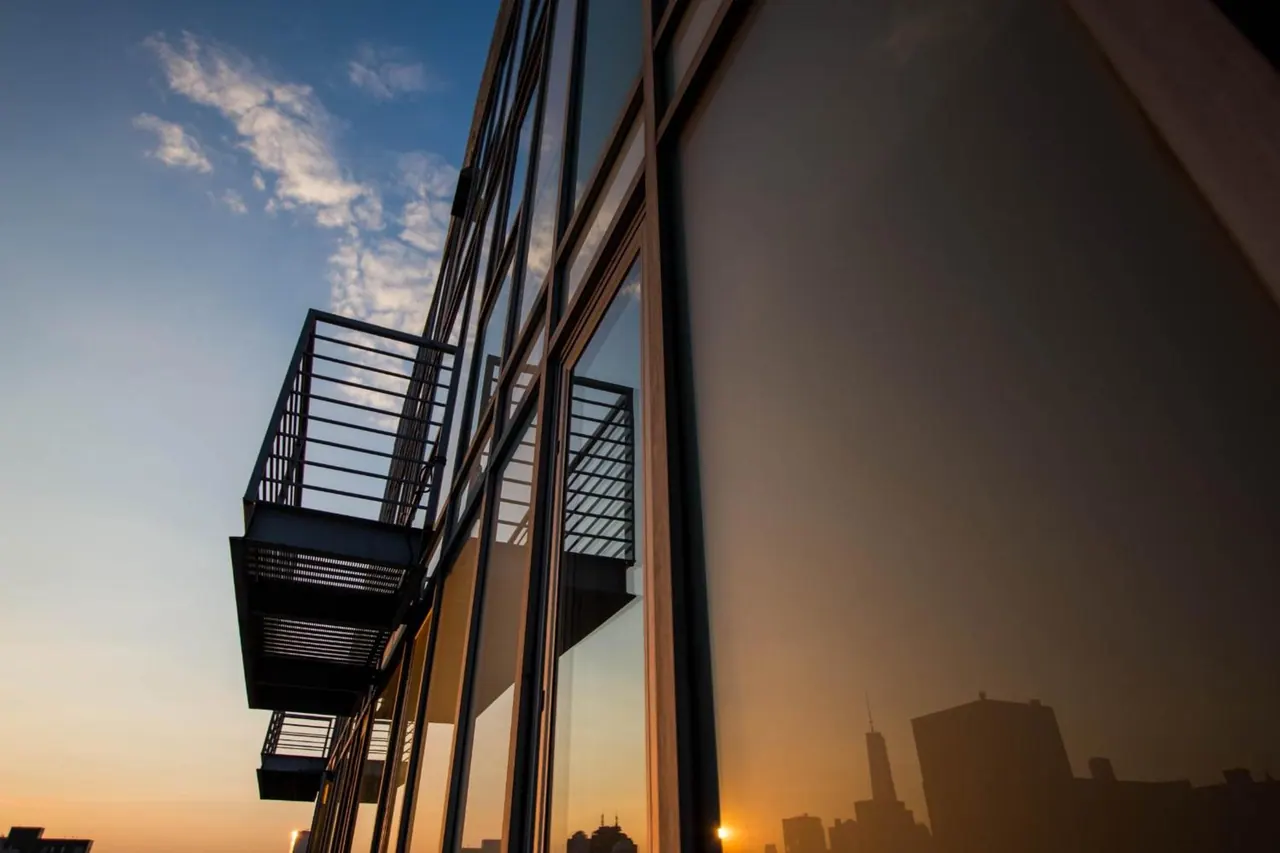 Exterior of a glass-fronted building with metal fire escape balconies, with a city skyline reflected in the windows at sunset.