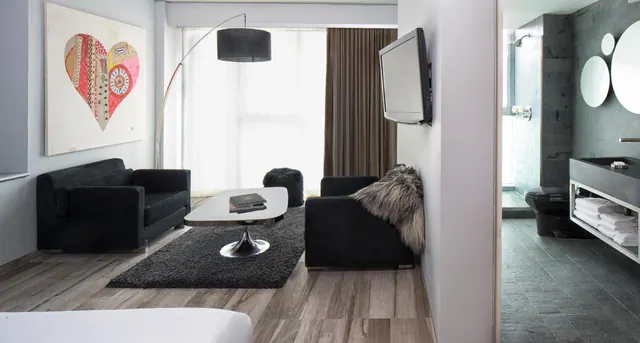 A modern hotel room with black seating, a coffee table on a dark rug, and a wall-mounted television, with a bathroom and walk-in shower visible on the right.