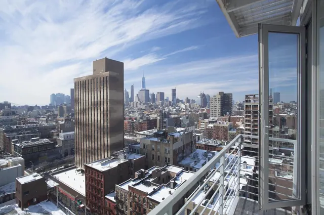 A view from a balcony over snowy rooftops and city buildings toward One World Trade Center under a partly cloudy sky.