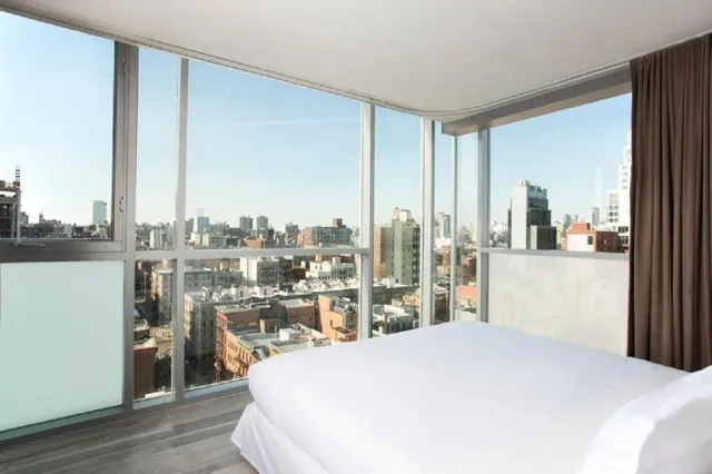 A bedroom with a made white bed facing floor-to-ceiling windows that overlook city buildings under a clear sky.