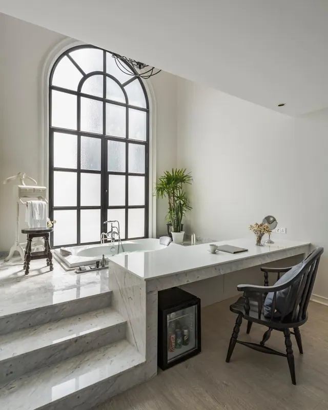 A marble-clad room with a bathtub on a raised platform beneath a tall arched window, with a desk, chair and small fridge to the right.