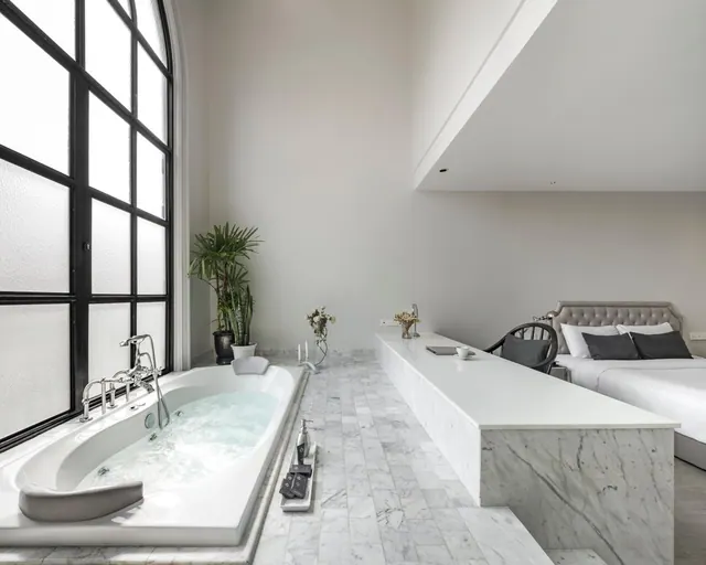 A bathtub filled with water sits beside a tall arched window, with a bed and a marble counter in the same room.