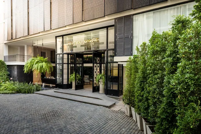 The entrance of Hotel Once has open glass doors, a hanging fern, and tall hedges along a cobbled driveway.