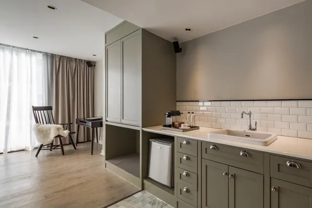 A corner of a hotel room with grey cabinets, a sink, a coffee machine, a mini fridge, a small desk and a chair with a sheepskin by a window with sheer curtains.