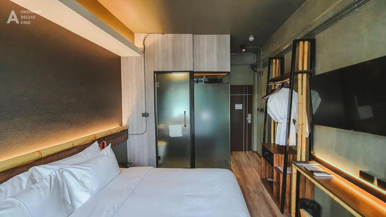 A bedroom with a king bed, a wall-mounted television, a work desk and an open wardrobe holding a bathrobe, with a frosted glass bathroom enclosure beside the bed.