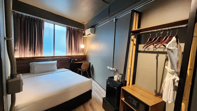 A small hotel bedroom with a double bed, a window with curtains, a desk and chair, a wall air conditioner, a kettle and fridge, and a wardrobe with a hanging bathrobe.