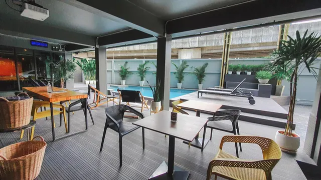 A covered outdoor dining area with wooden and dark tables and yellow chairs, with a swimming pool, sun loungers and potted plants beyond.