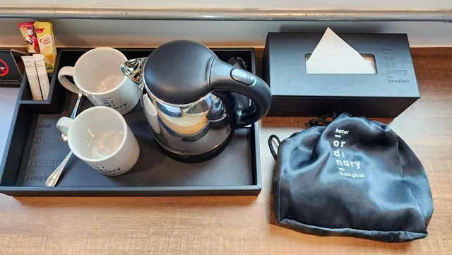 A black tray holds a steel kettle, two white mugs, a spoon and creamer sachets beside a black tissue box and a black drawstring pouch on a wooden surface.
