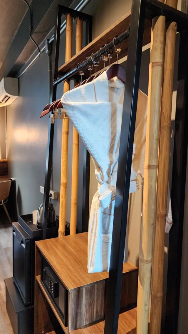A corner of the room shows a metal and bamboo clothing rack with white bathrobes on wooden hangers, a kettle, a minibar fridge and a safe in a wooden cabinet, with a wall air conditioner above.
