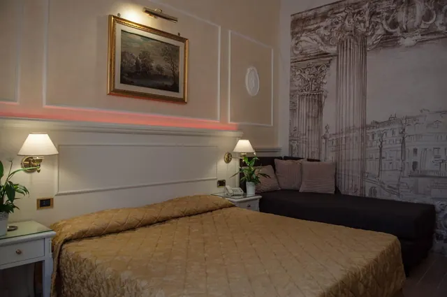 A double bed with a gold quilted cover sits beside bedside tables with lamps and plants, with a dark sofa under a wall mural of classical columns and buildings.