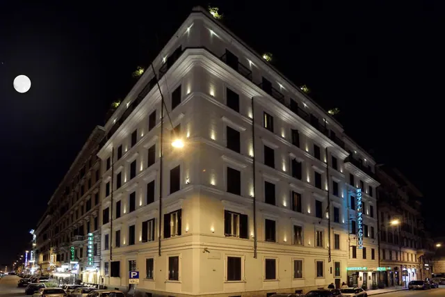 Night exterior of a corner hotel building with uplit white facade, a vertical blue sign reading Hotel Palladium, parked cars along the street and a moon in the dark sky.