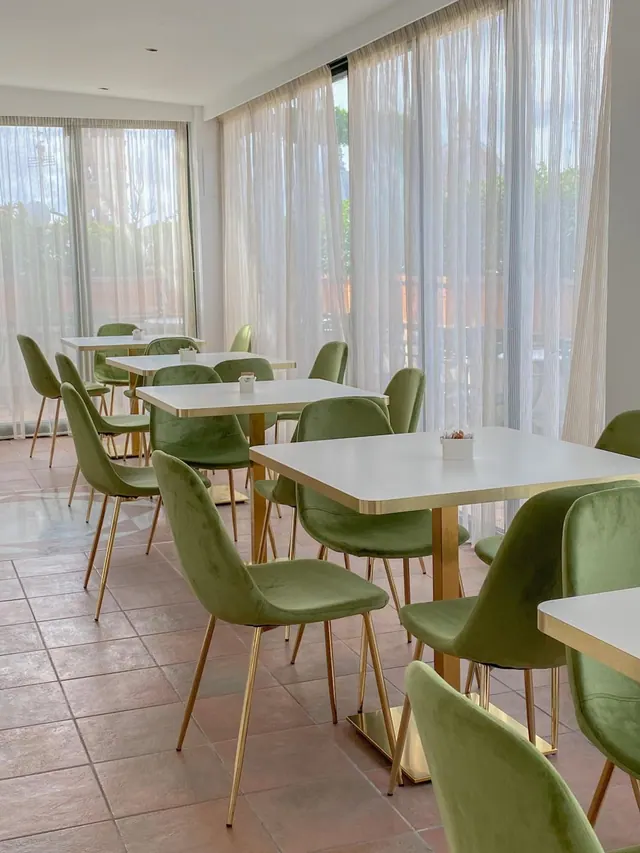 A dining room with green velvet chairs and white tables with gold edges, set beside large windows with sheer curtains.