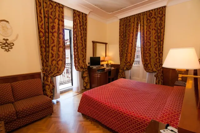 A classic hotel bedroom with a red patterned double bed, damask curtains, a two-seat sofa, a desk with a computer monitor, and an open door to a small balcony.