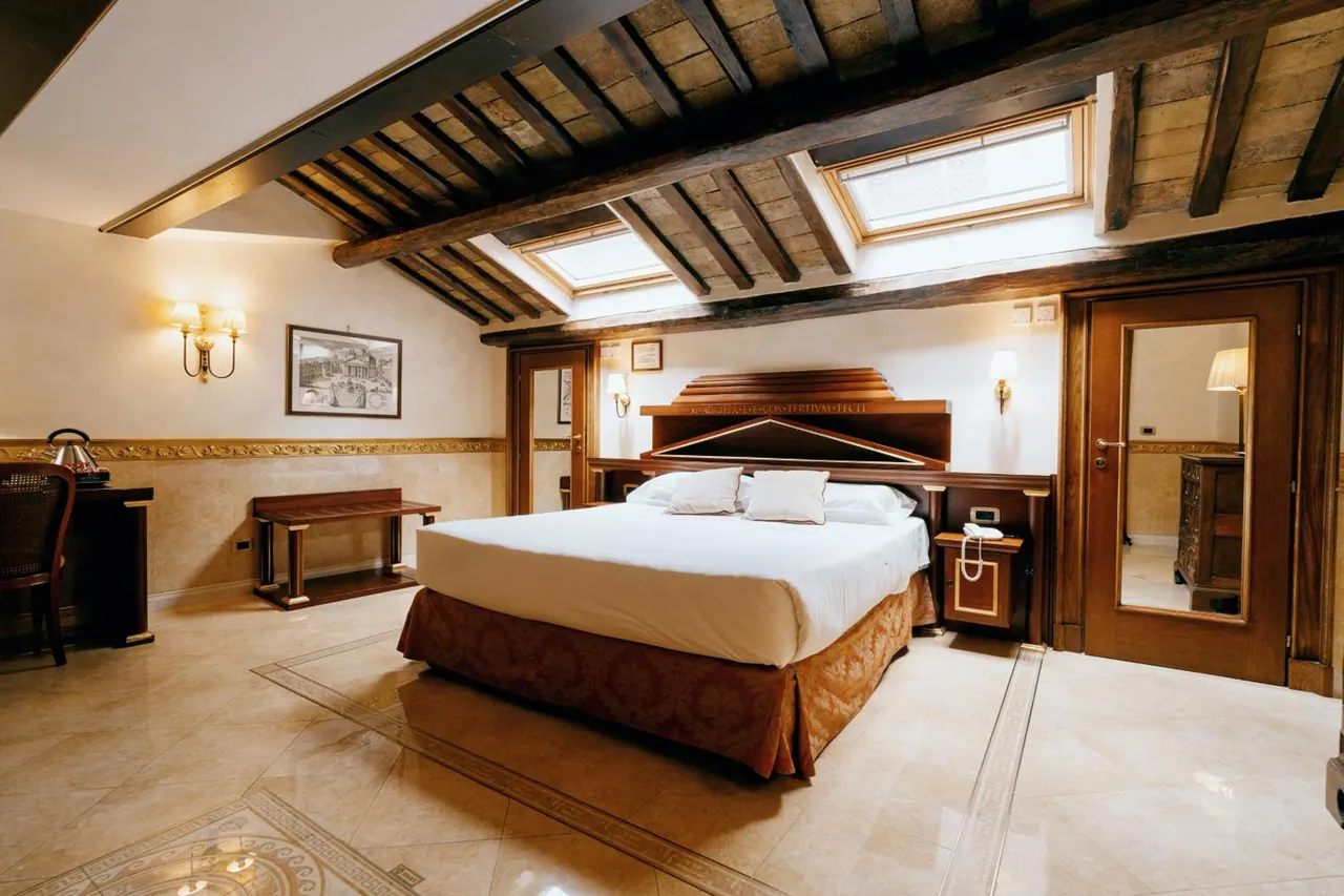 A bedroom with a double bed under exposed wooden beams and skylights, with a desk and chair on the left and a wooden door on the right.