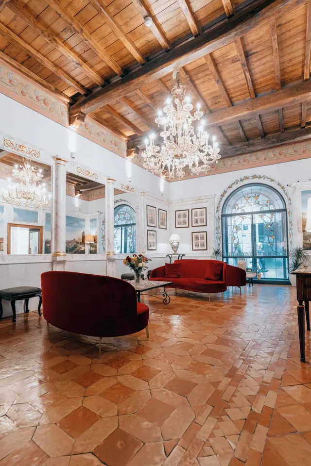 A lounge with red velvet sofas, a glass chandelier, a beamed wooden ceiling and a terracotta tiled floor, with an arched stained-glass window at the far end.