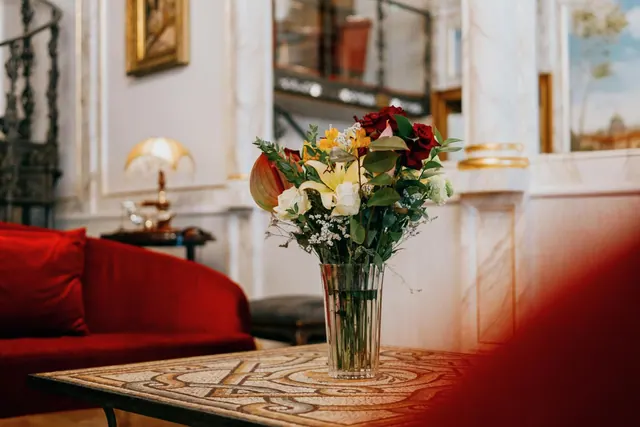 A vase of mixed flowers stands on a mosaic-topped table in front of a red sofa, with a table lamp and a gilded framed painting in the background.