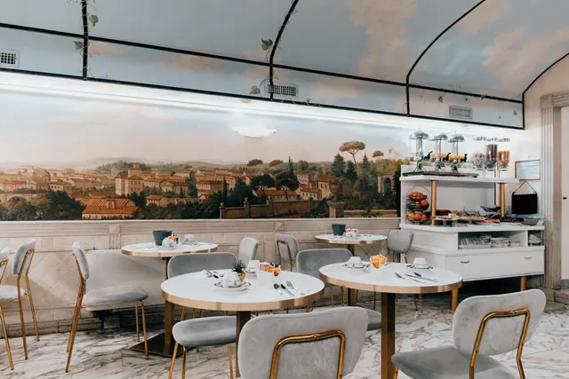 A breakfast room with round white tables, grey velvet chairs and a buffet station beneath a painted wall mural of a hilly town landscape.