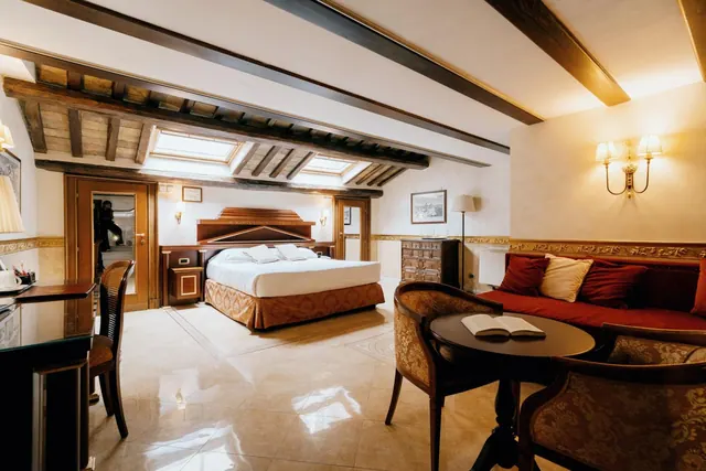 A bedroom with a double bed, a red sofa and armchairs around a round table, under exposed wooden beams and skylights.