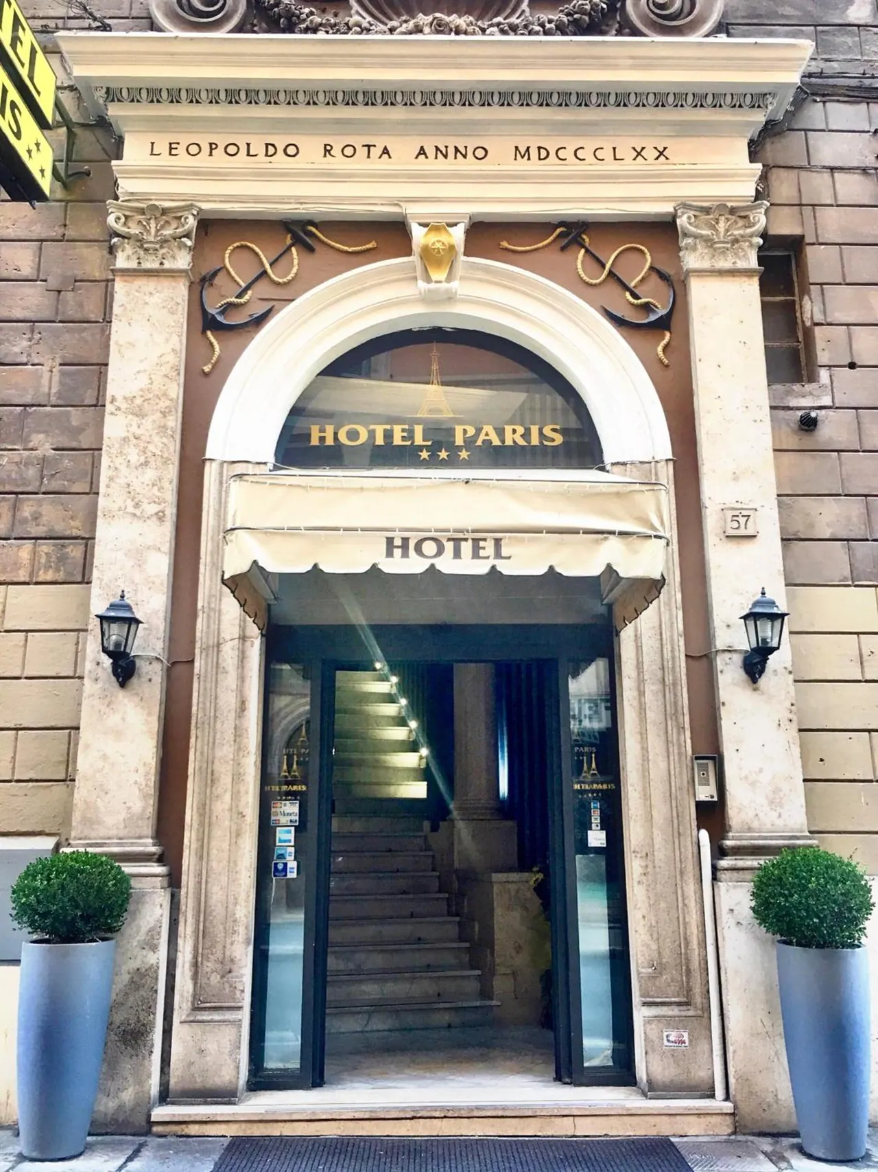 Front entrance of Hotel Paris, with an arched doorway, a cream awning reading HOTEL, a carved inscription above, and potted topiary on either side.