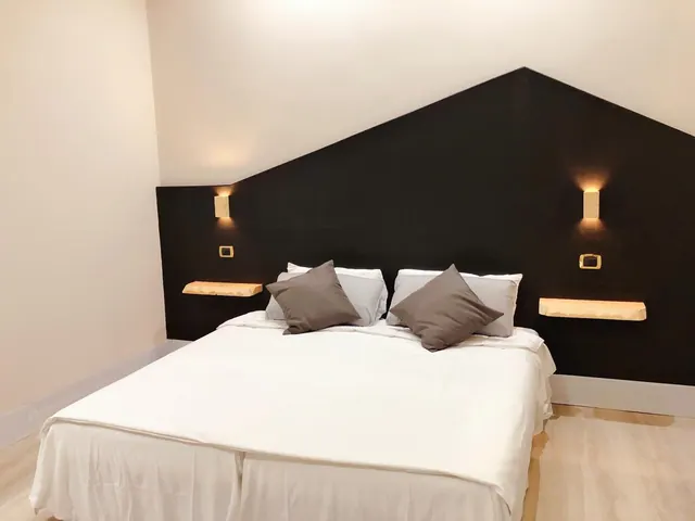 A double bed with white linen and grey cushions stands against a black angled accent wall with wall lights and wooden floating shelves.