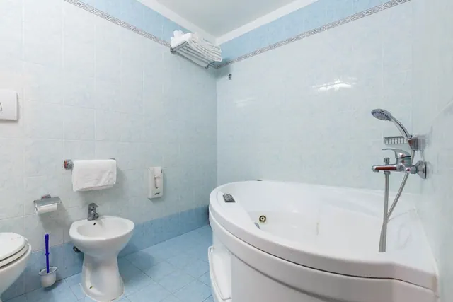 A blue-tiled bathroom with a corner bathtub, handheld shower, bidet, and toilet, with folded towels on a rack above.