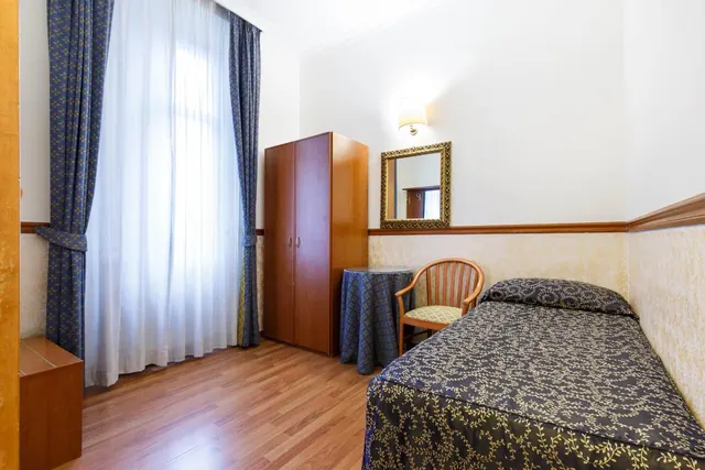 A bedroom with a single bed, a wooden wardrobe, a chair beside a round table, a wall mirror and a tall window with sheer and blue curtains.