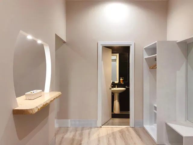 An empty room with a live-edge wooden shelf and recessed lights on the left, a white wardrobe on the right, and an open door to a bathroom with a pedestal sink.