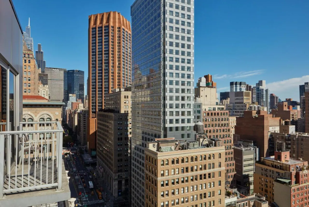 A view over dense midtown skyscrapers and older brick buildings, taken from a balcony with a white metal railing in the foreground.