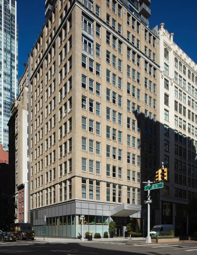Exterior of a tall beige brick hotel building with a glass ground floor and a canopy reading Hotel Park Ave, beside a street corner with a 30 St sign.
