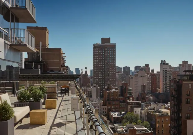 A rooftop terrace with cushioned seating, planters and string lights overlooks a dense city skyline of mid-rise and high-rise buildings under a clear sky.
