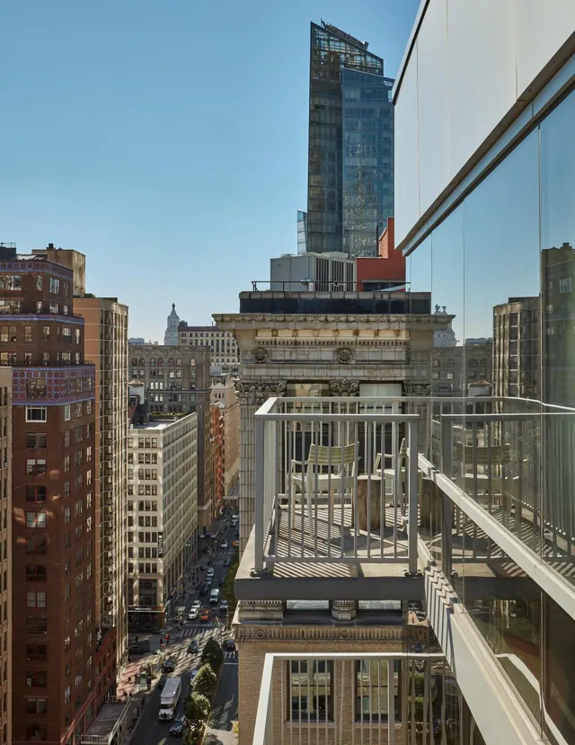 A railed balcony with two chairs overlooks a city street lined with tall buildings and traffic under a clear blue sky.
