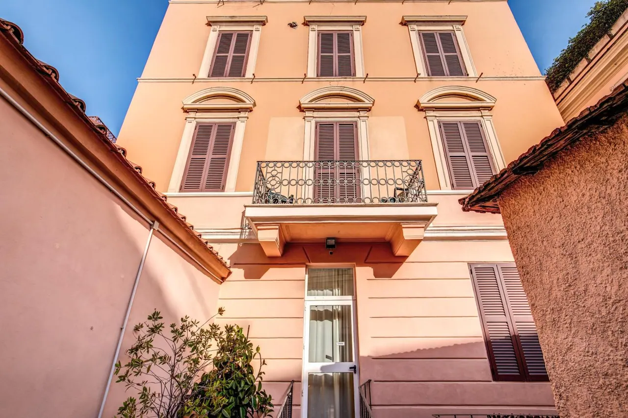 Looking up at a peach-coloured building facade with shuttered windows, a wrought iron balcony, and a glass entrance door at street level, under a clear blue sky.