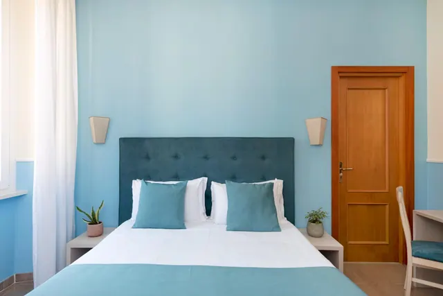 A bedroom with a double bed under a tufted teal headboard, blue walls, a wooden door, and a desk chair at the right edge.