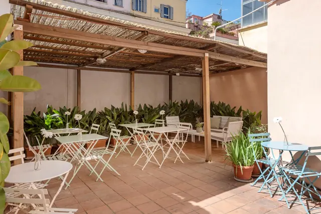 A covered terrace with a reed-roofed wooden pergola, white and blue bistro tables and chairs, potted plants and an outdoor sofa with cushions.