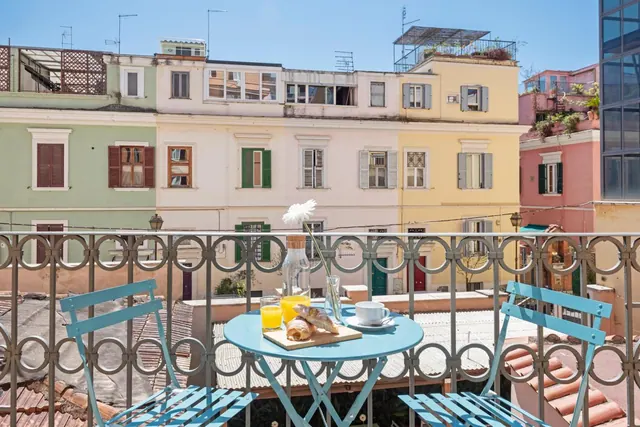 A small blue bistro table with two chairs, orange juice, croissants and a coffee cup sits on a balcony overlooking colourful multi-storey buildings.