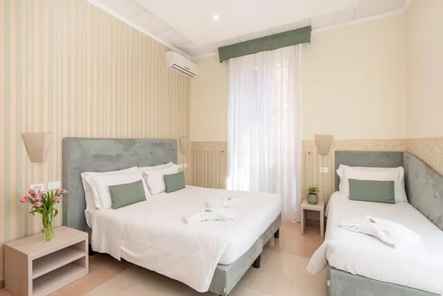 A bedroom with a double bed and a single bed, striped wallpaper, a wall-mounted air conditioner and a window with white curtains.